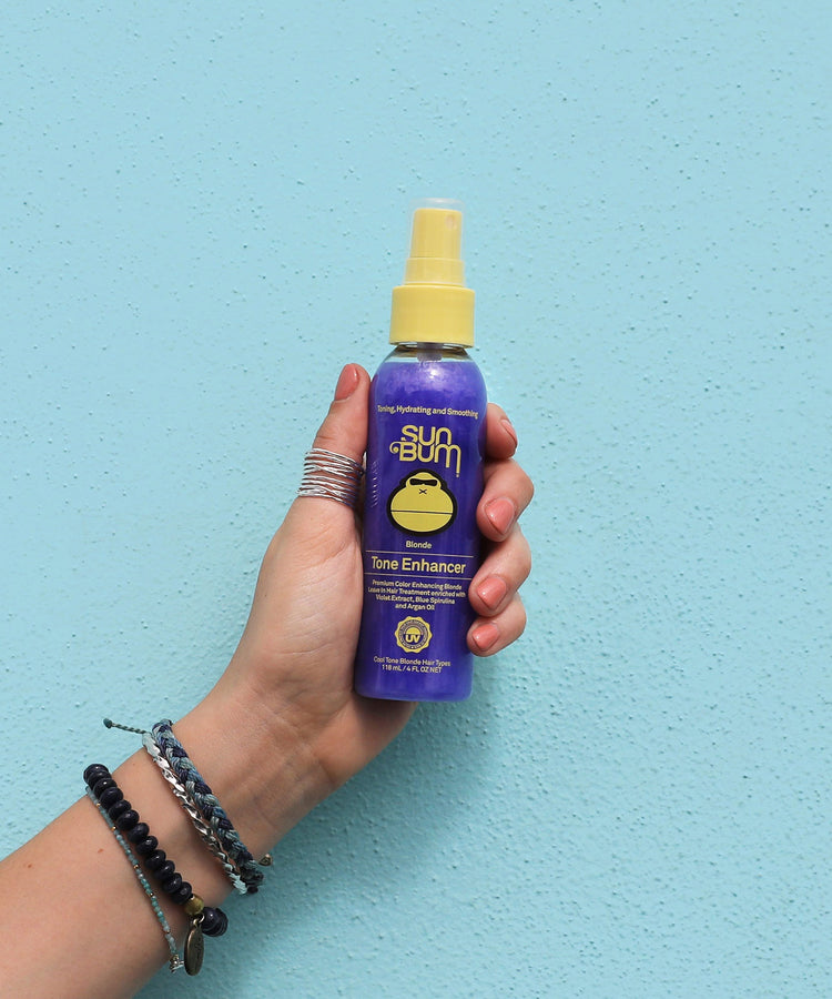 Hand holding a purple bottle of Sun Bum Turbo Enhancer against a blue background