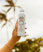 Hand holding a Sun Bum sunscreen spray bottle with a blurred natural background