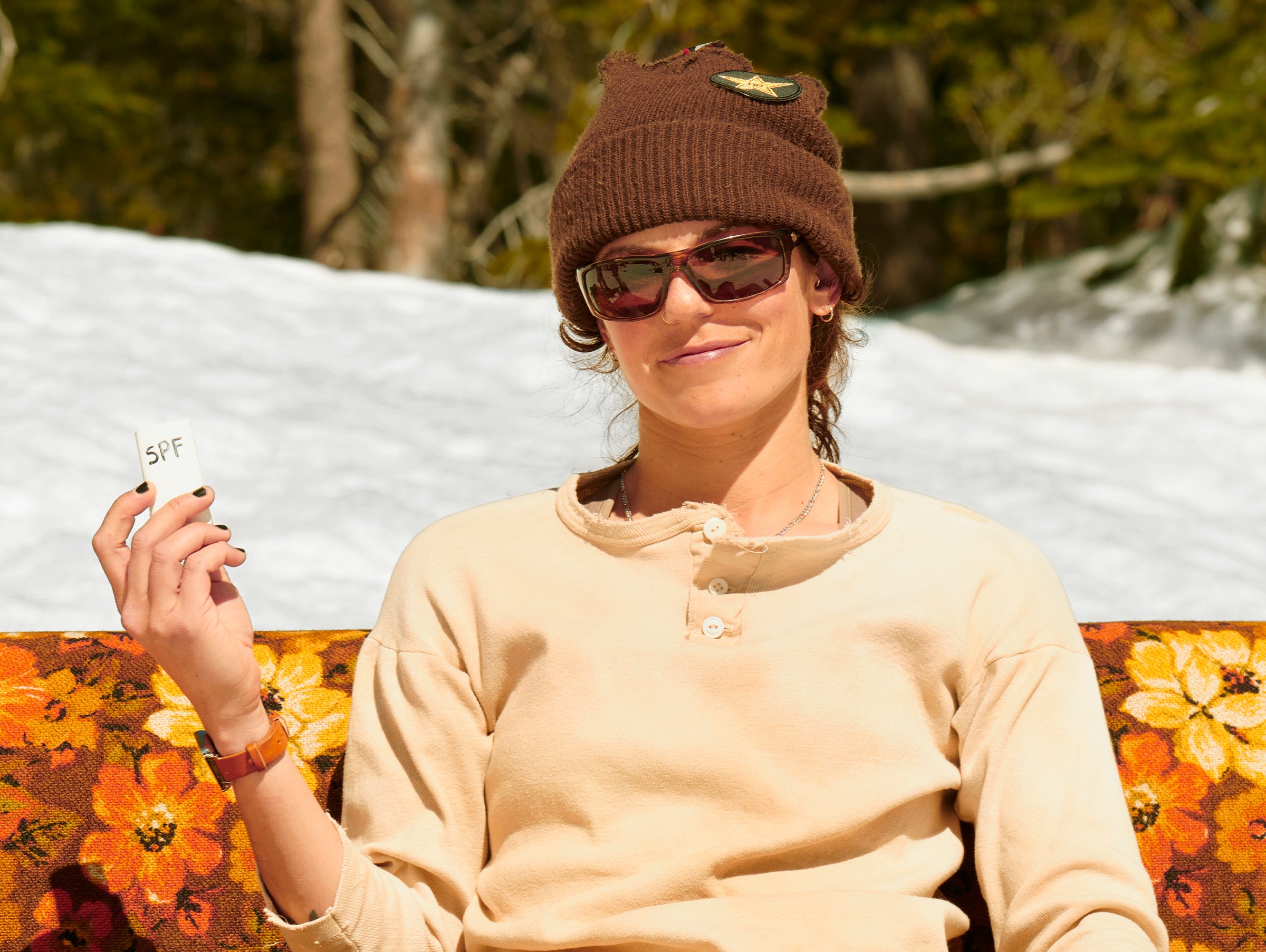 Person wearing a brown beanie and sunglasses, sitting on a floral-patterned bench with snow and trees in the background.