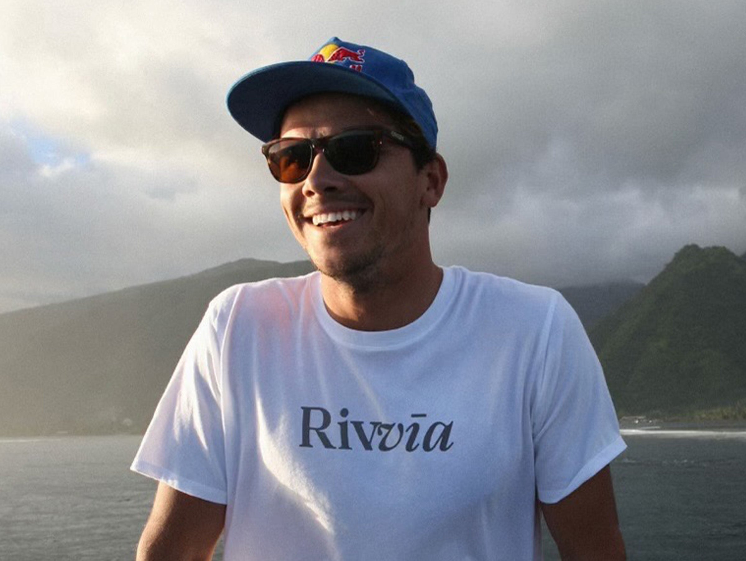 Man wearing a white t-shirt with 'Rivvia' logo and a blue cap with a red bull logo, standing in front of a mountainous landscape.
