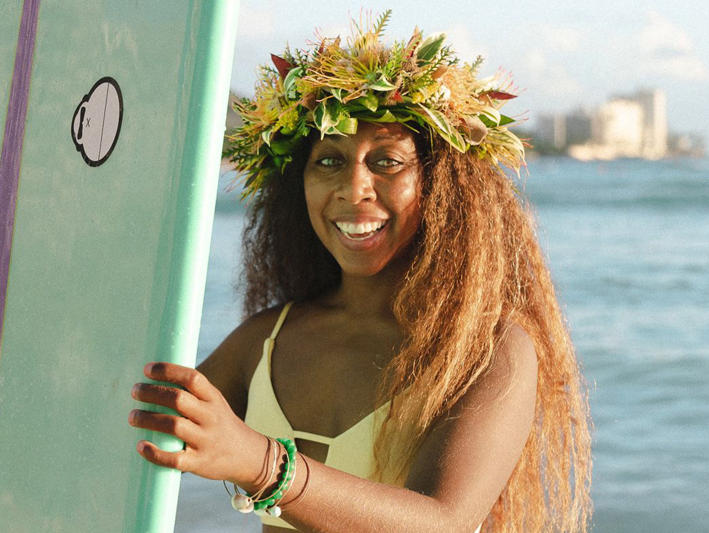 Woman with a floral headpiece holding a surfboard by the water