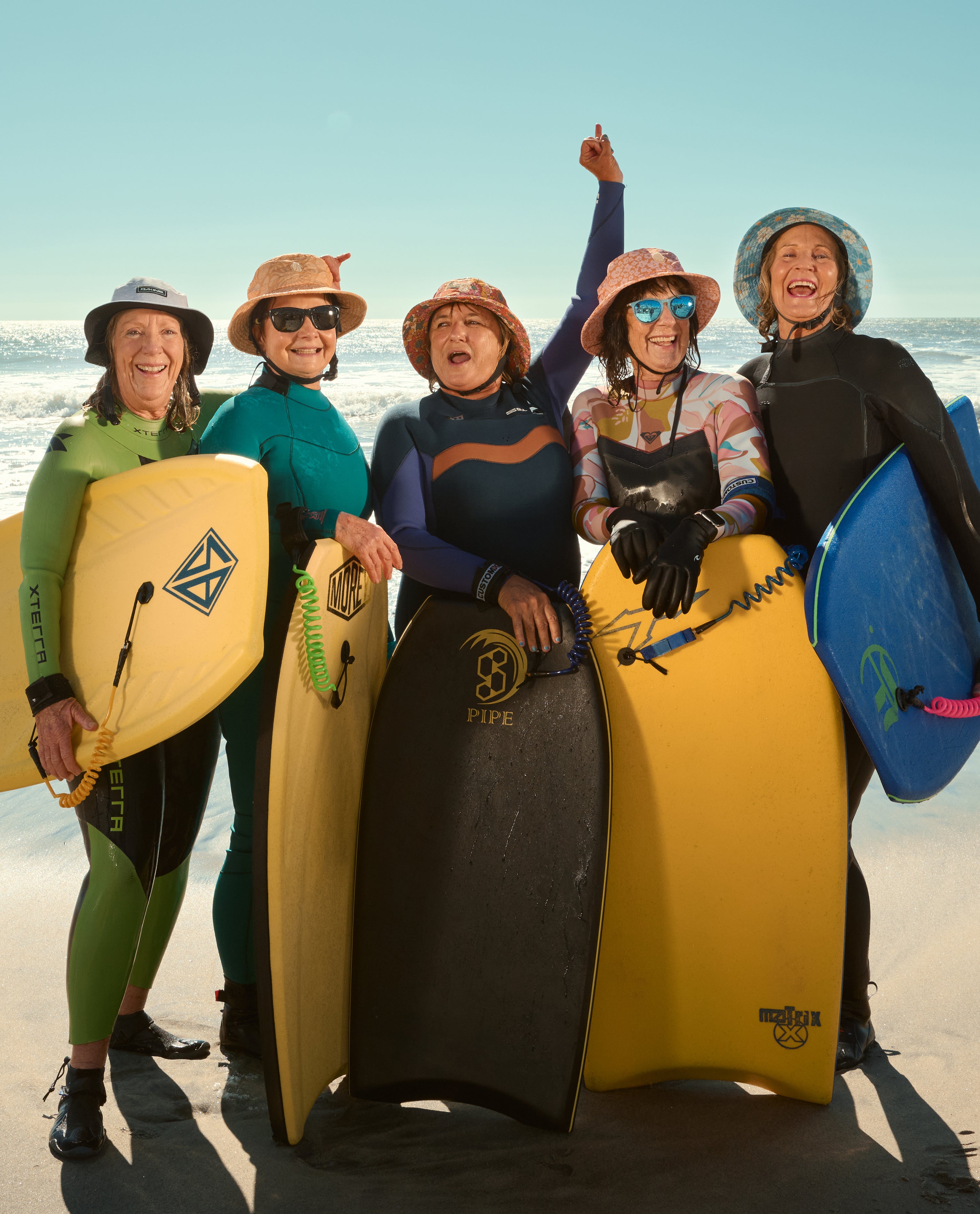 Five people on a beach holding bodyboards with a clear sky and ocean in the background