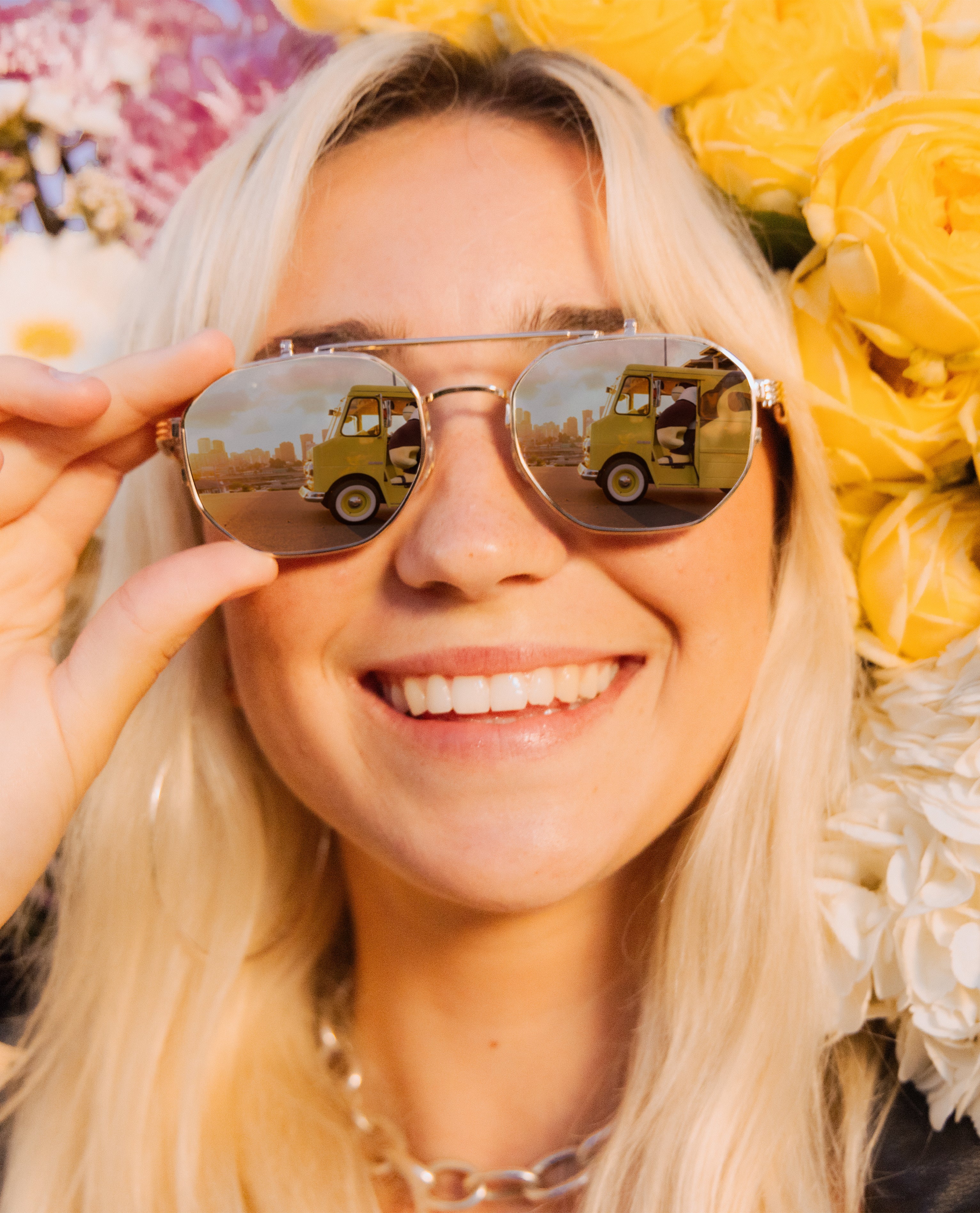 Woman wearing sunglasses with a reflection of a yellow truck, surrounded by flowers.