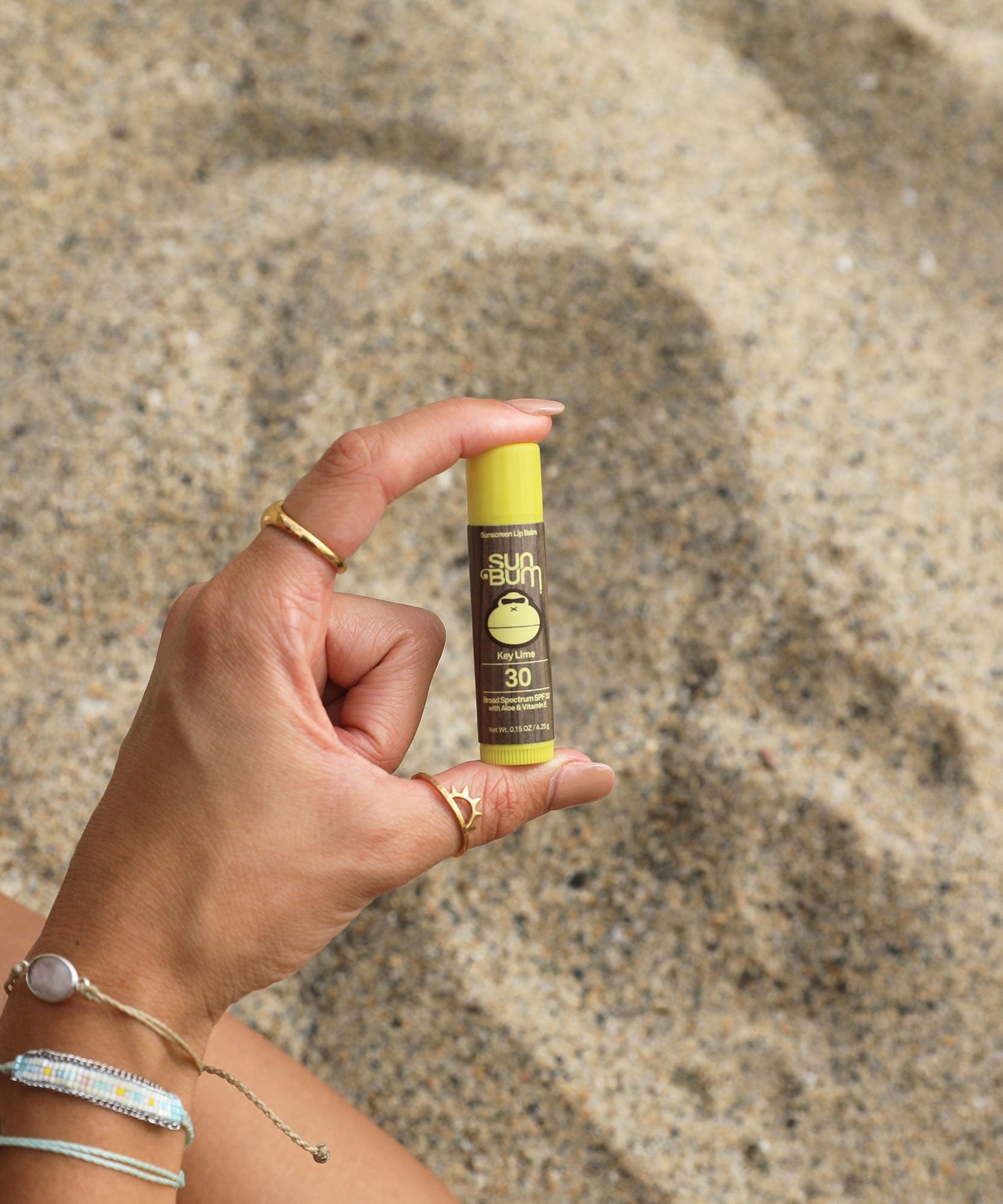 Hand holding a Sun Bum sunscreen stick against a sandy background