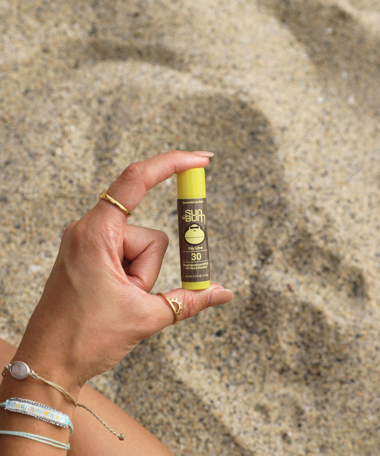 Hand holding a Sun Bum sunscreen stick against a sandy background