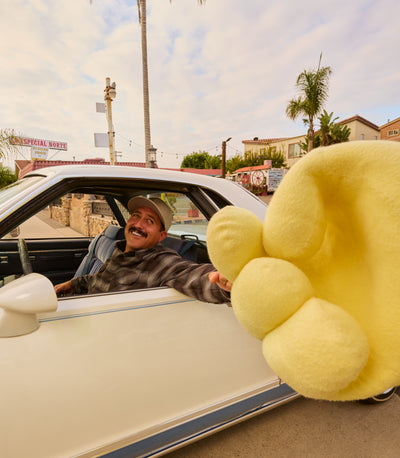 Man sitting in a car with a large yellow plush toy in the foreground, palm trees and buildings in the background.