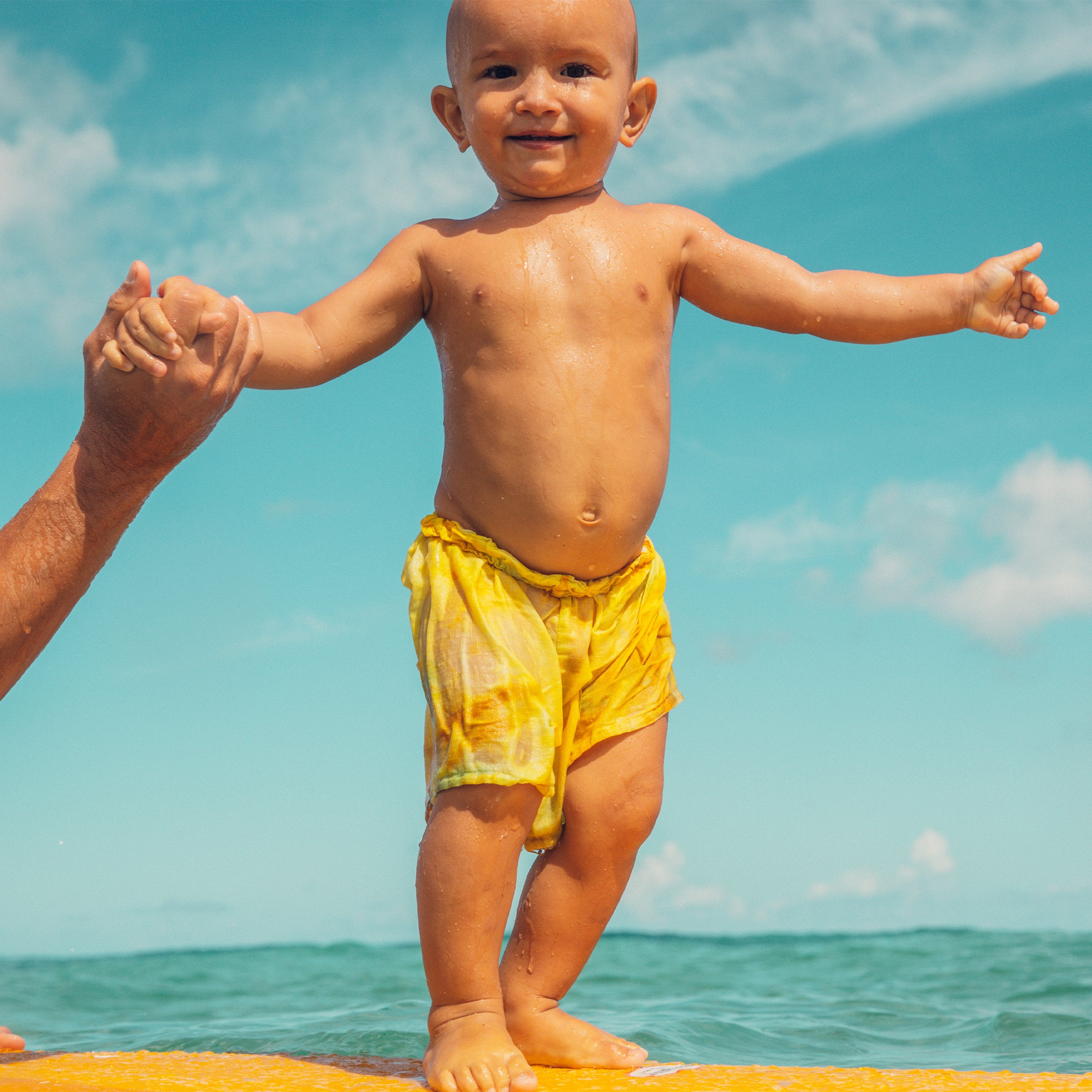 Child in yellow swim shorts standing on a surfboard with ocean and sky in the background