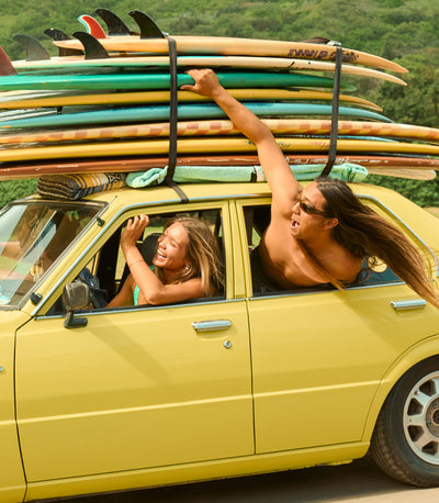 Two people in a yellow car with surfboards on top, surrounded by greenery.