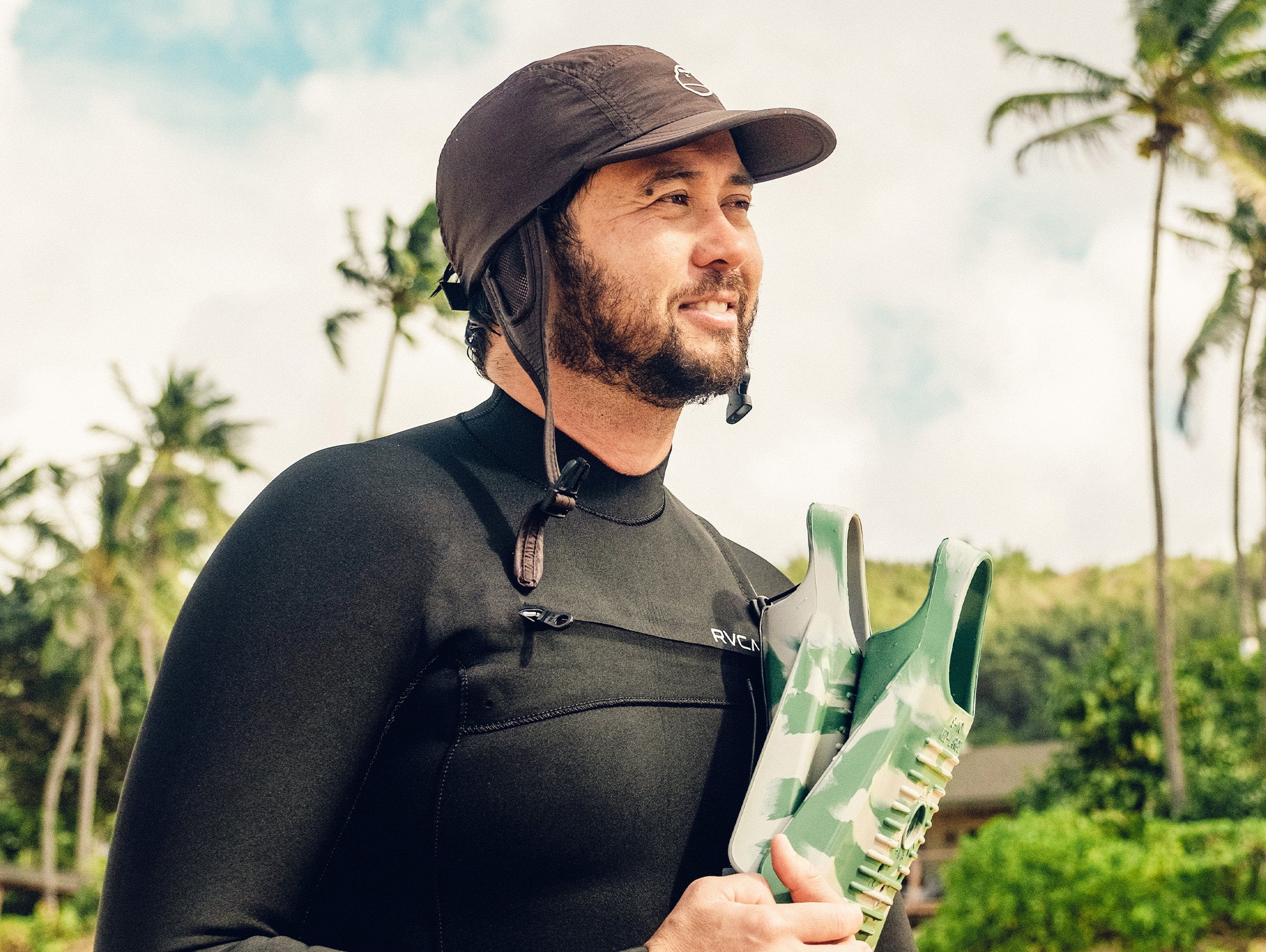 Man in a wetsuit holding a green surfboard with palm trees in the background